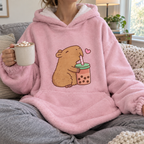 Adult wearing the pink capybara blanket hoodie on a sofa with a mug