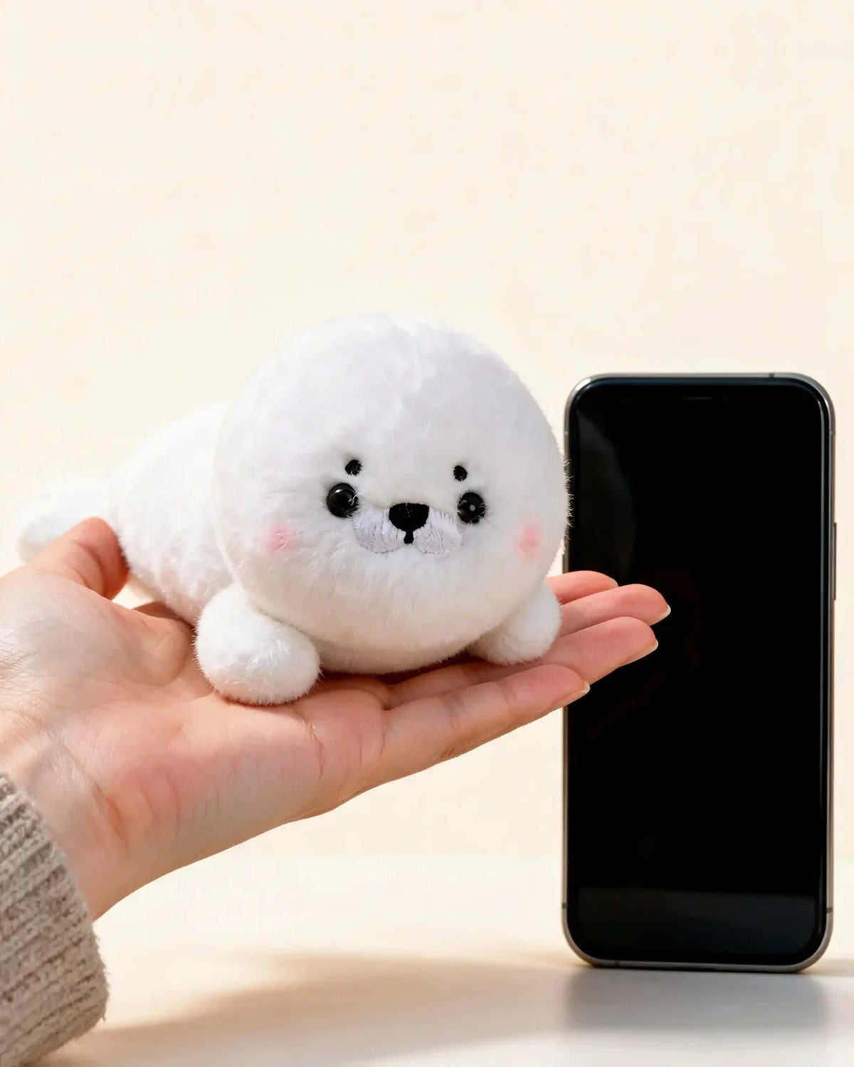 Floe, the Sea Friends seal plush, resting on a hand next to a smartphone to show its size