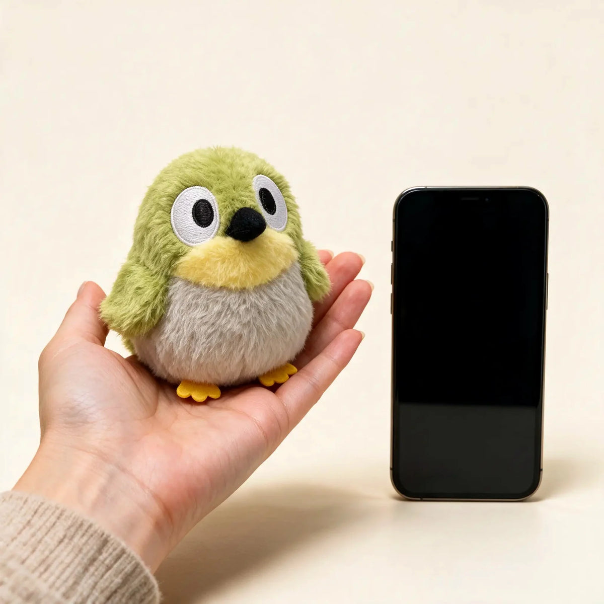 Mossy, the green Feather Friends bird plush, sitting in a palm next to a smartphone to show its size
