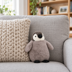 Gray penguin chick plush standing on a sofa beside a knit pillow
