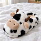Close-up of a spotted cow plush slipper on white linen bedding showing its 3D cow face and fur